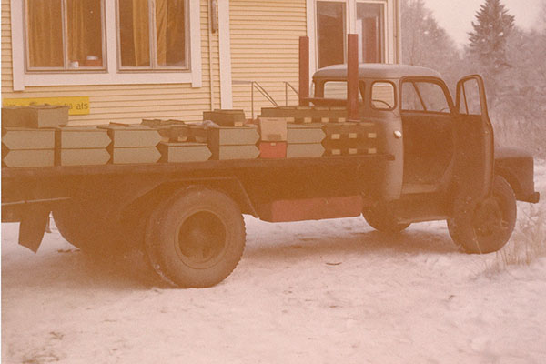 Bedford-kuorma-auto lastattuna laatikoilla talvisessa pihapiirissä keltaisen puutalon edessä.