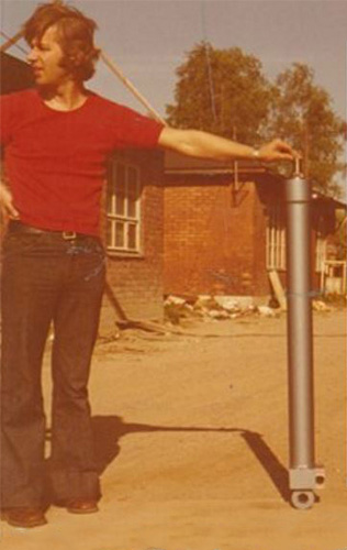 Nuori mies esittelee hydraulisylinteriä tiilirakennuksen pihalla 1970-luvulla.