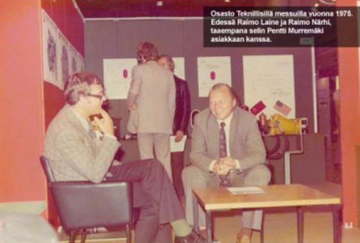 "Teknillisten messujen osasto vuonna 1975. Etualalla kaksi miestä, Raimo Laine ja Raimo Närhi, istuvat keskustelemassa pöydän ääressä. Taustalla Pentti Murremäki seisoo selin asiakkaan kanssa. Seinillä on julisteita ja teknisiä piirustuksia